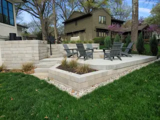 Raised concrete patio with block retaining walls, integrated planter of ornamental grasses, Adirondack chairs around a fire pit, and white stone edging